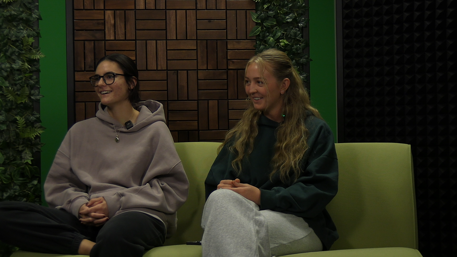 Leah Wolf and Ruby Hladek sit on a green couch smiling at an interviewer off-camera